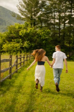 Couple holding hands running through green meadow by wooden fence with mountains and trees in background.
