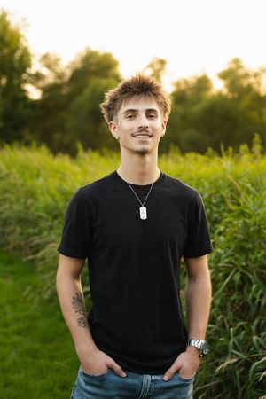 Young man with mustache in black t-shirt and dog tag necklace smiling outdoors in green field at sunset.