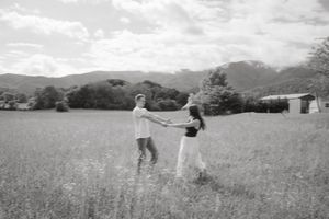 Black and white photo of a couple holding hands dancing in a scenic meadow with mountains in the background.