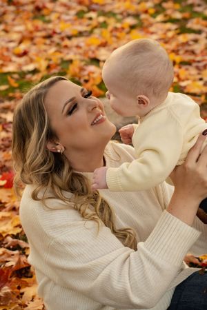 A smiling mother in a cream sweater holds her baby outdoors among colorful autumn leaves in a park.