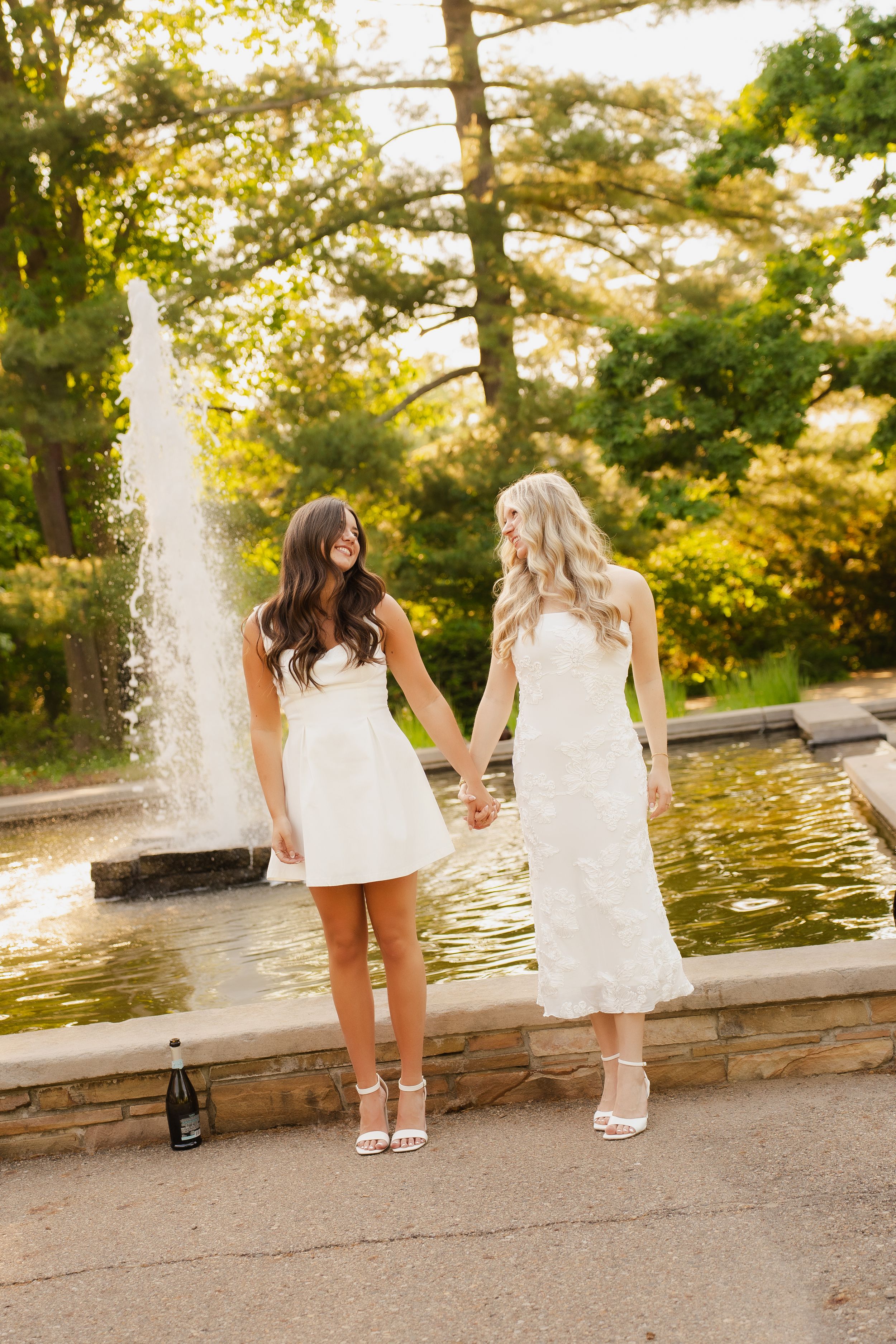 Two brides in white dresses hold hands by a fountain in a lush garden setting on their wedding day.