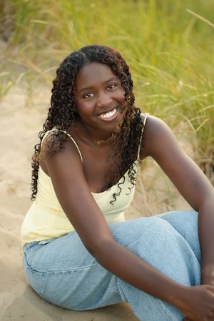 Smiling young Black woman with curly hair sitting on sandy beach in yellow top and jeans.