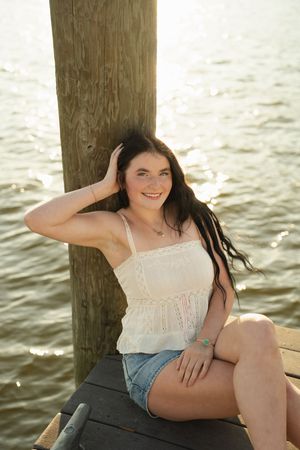 Young woman in white top and denim shorts sitting on a wooden dock by the water on a sunny day.