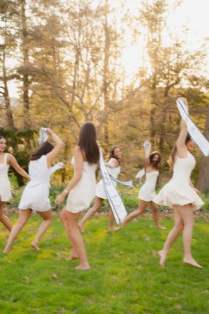 Group of women in white dresses dancing barefoot on green grass surrounded by autumn trees.