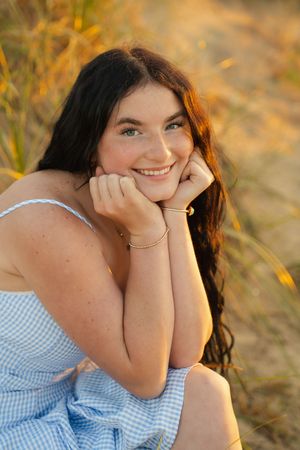 Young woman with dark hair smiling outdoors in blue gingham dress, resting chin on hands in golden field.
