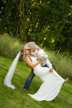 Bride and groom share a romantic dip kiss outdoors, she holds a bouquet in a white gown with veil, he wears a cowboy hat.
