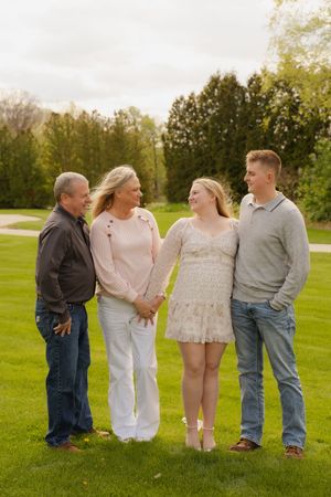 Family of four smiling and holding hands on a lush green lawn surrounded by trees.