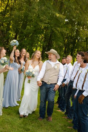A bride and groom in country-style attire walk hand-in-hand surrounded by bridesmaids in blue and groomsmen outdoors.