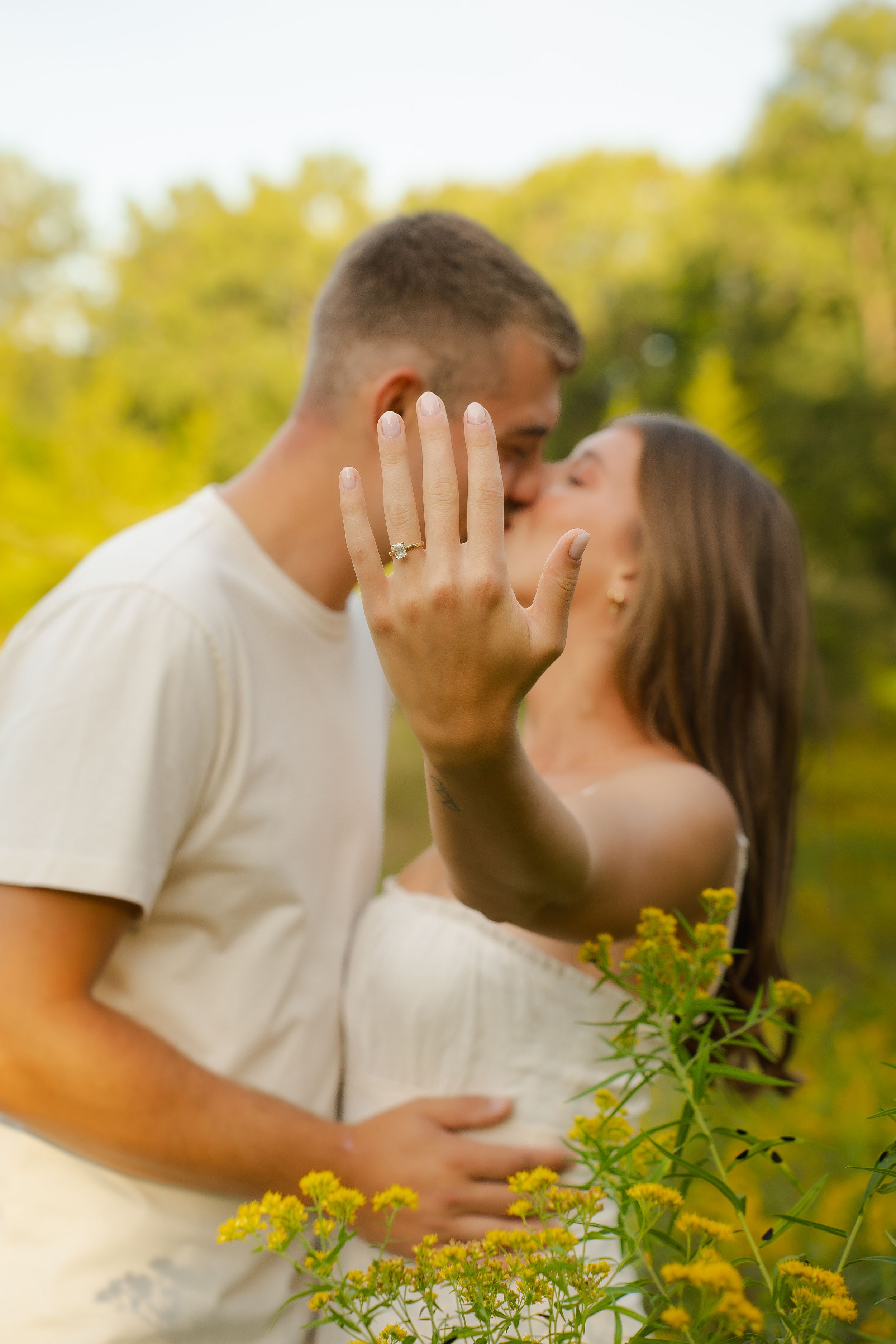 Couple kissing outdoors while showing off engagement ring, surrounded by yellow wildflowers and green trees.