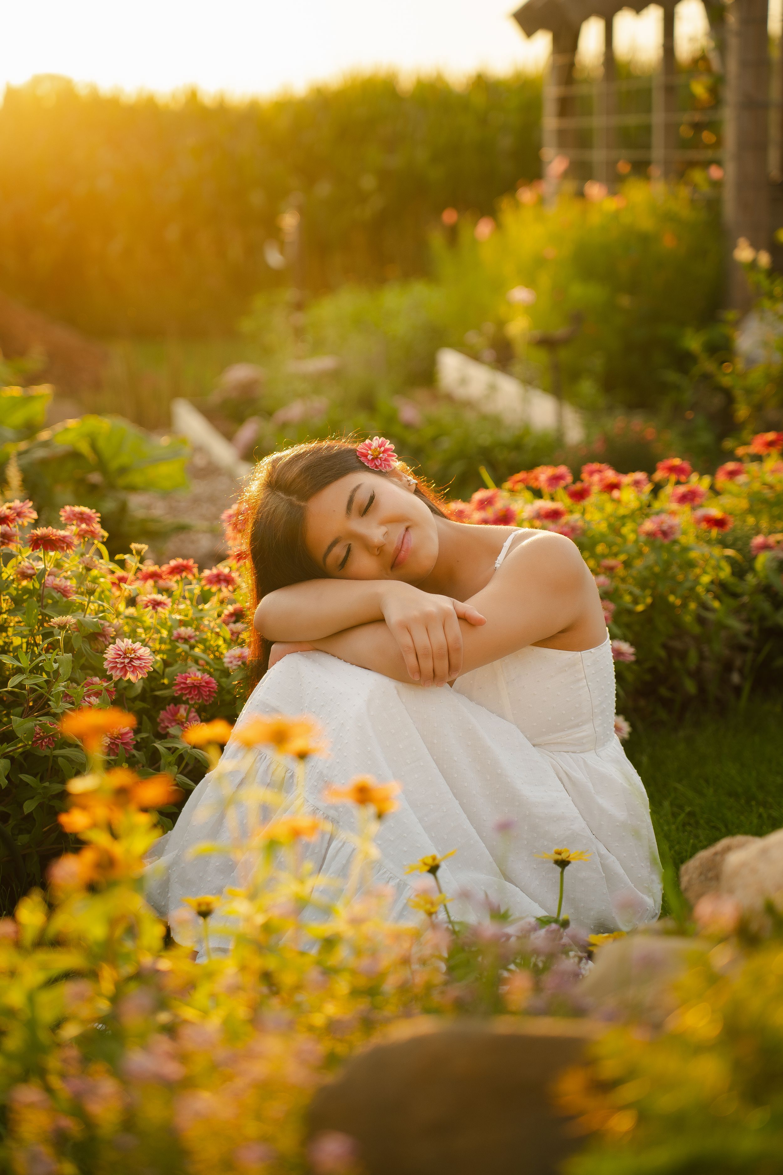 Young woman in white dress sitting peacefully among colorful garden flowers at golden sunset.
