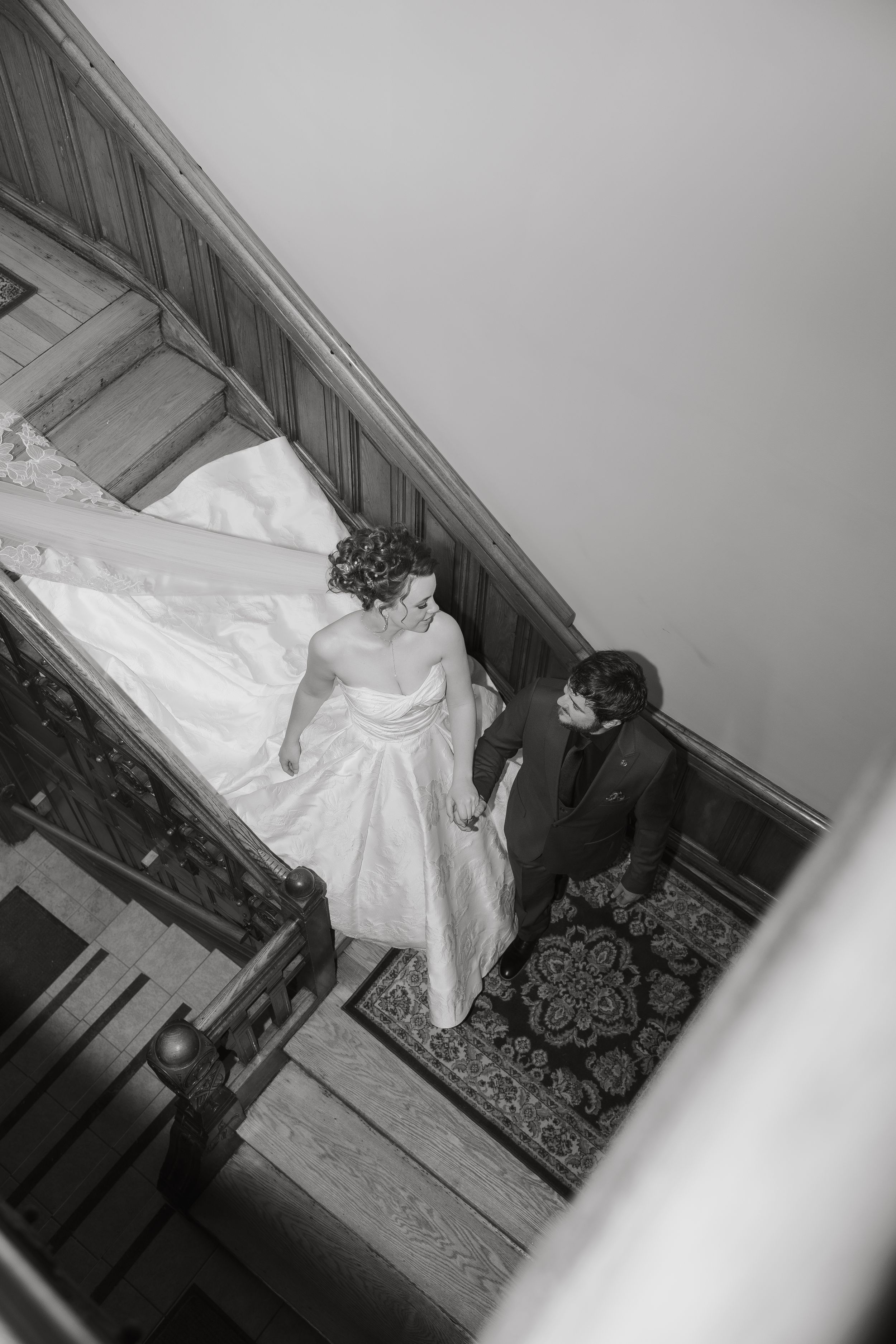 Black and white aerial view of bride and groom holding hands on ornate carpeted staircase, bride in flowing white gown.