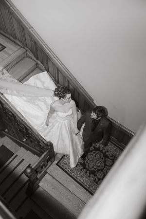 Black and white aerial view of bride and groom holding hands on ornate carpeted staircase, bride in flowing white gown.