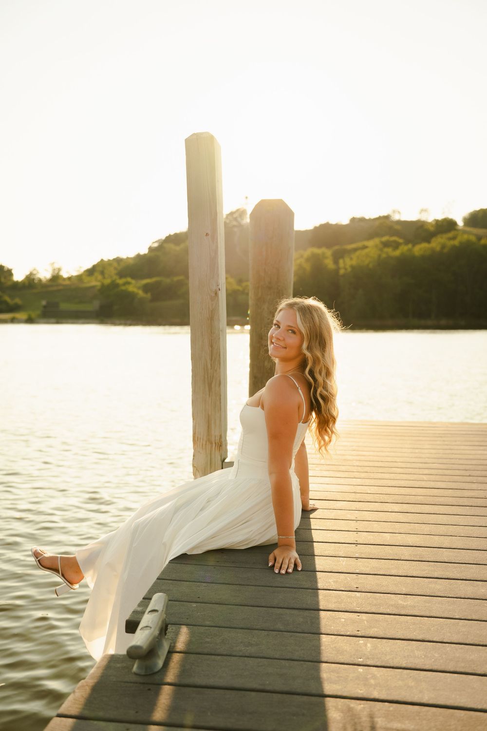 Woman in white dress sitting on wooden dock at sunset by calm lake with lush green trees in background.