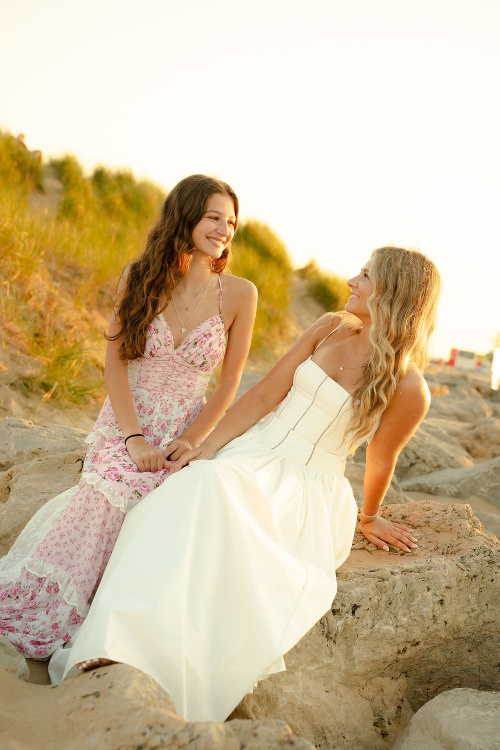 Two young women in elegant dresses sitting on sandy beach dunes at golden hour, smiling and enjoying each other's company.