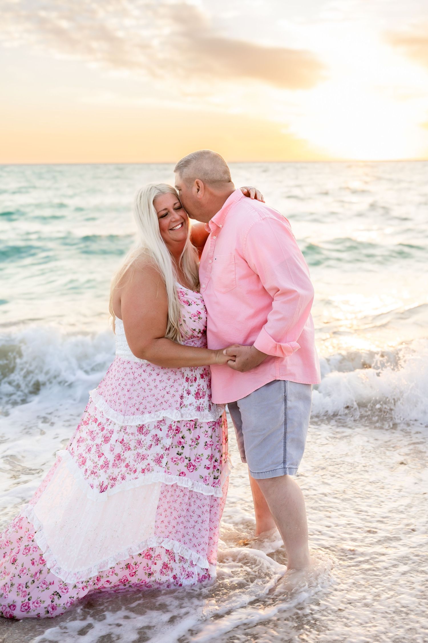 Romantic couple embracing in ocean waves at sunset, woman in pink floral maxi dress and man in pink shirt.