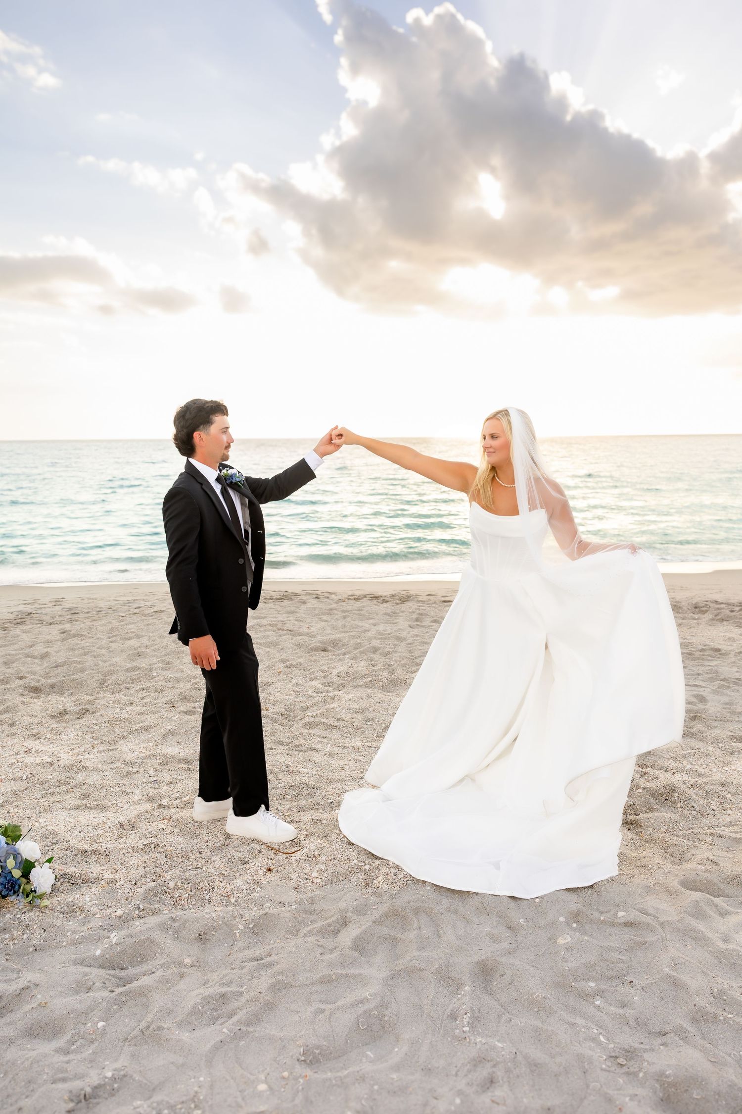 Bride and groom dancing on a sandy beach at sunset, groom in black suit spinning bride in white gown.