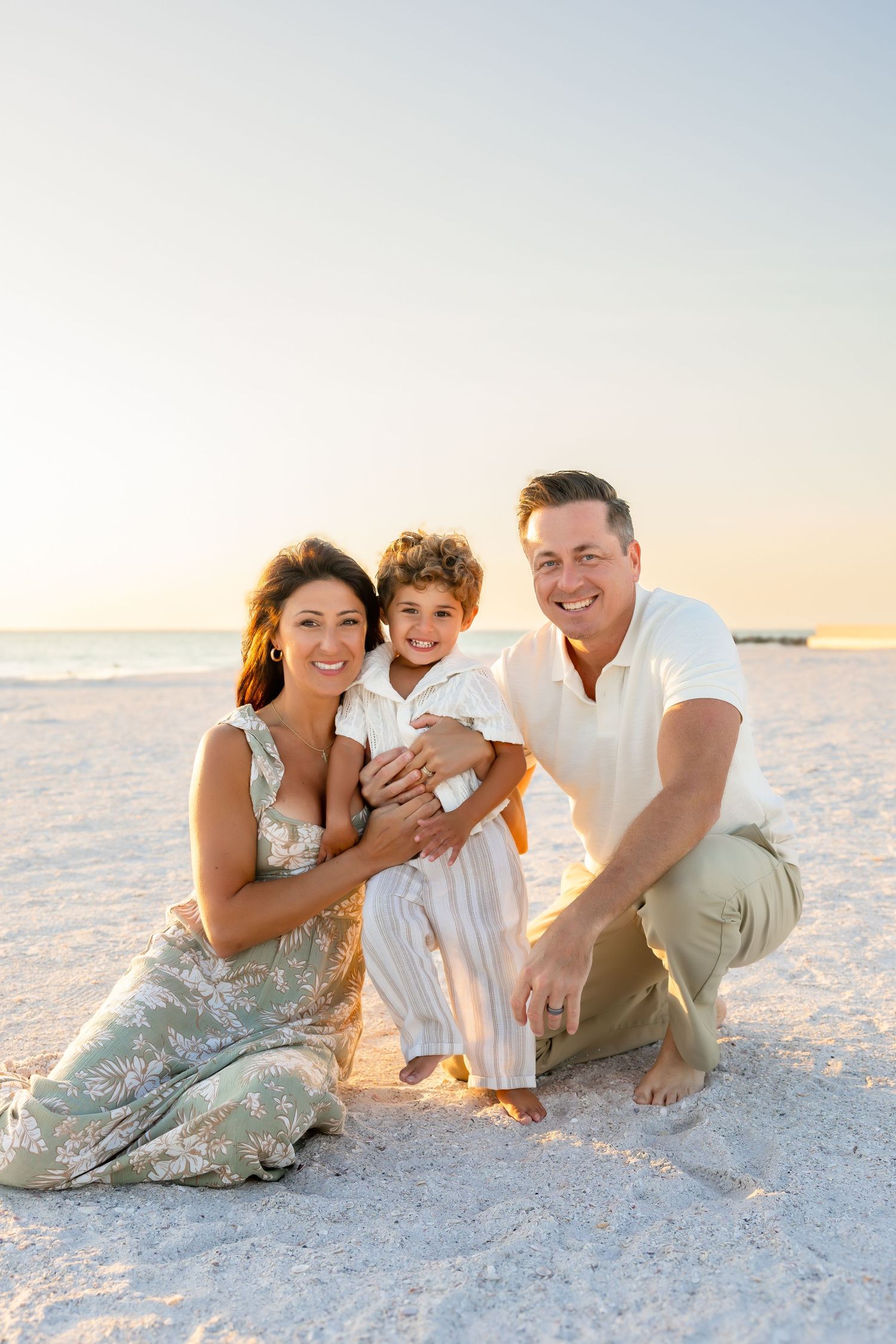 Happy family of three sitting on a white sand beach at golden hour, parents smiling with young child.
