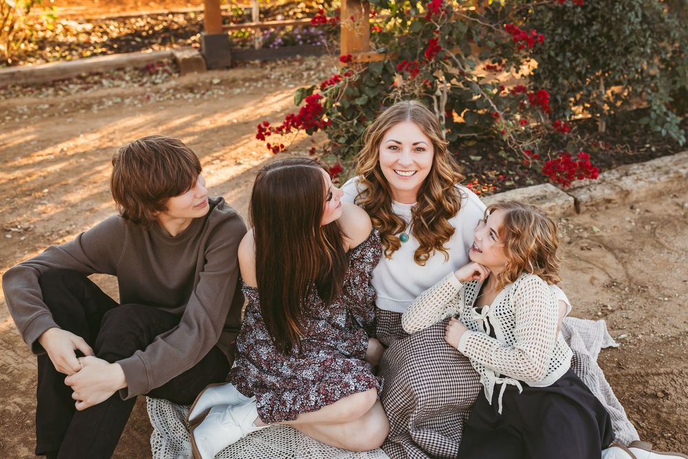Four siblings laughing together outdoors at sunset near red flowering bushes during a family photo session.