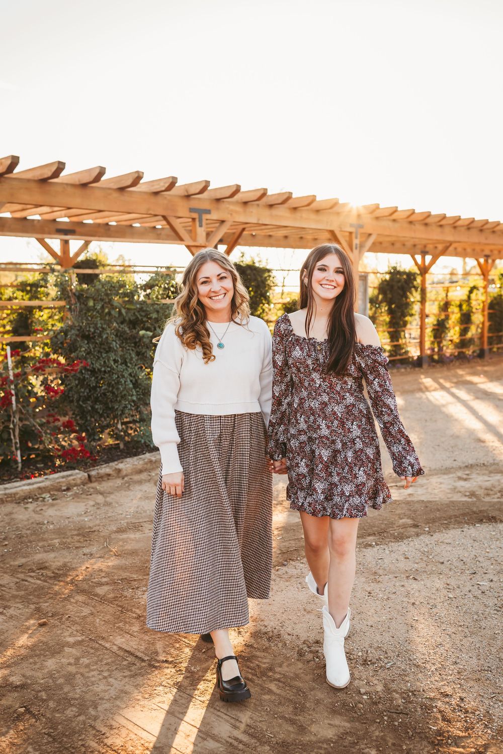 Two smiling women walk arm-in-arm in floral and knit outfits under a wooden pergola at golden hour.