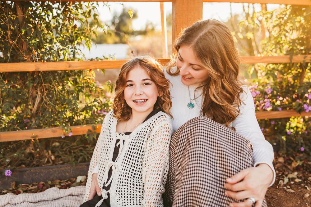Two smiling girls pose together outdoors near a wooden fence surrounded by greenery in warm sunlight.