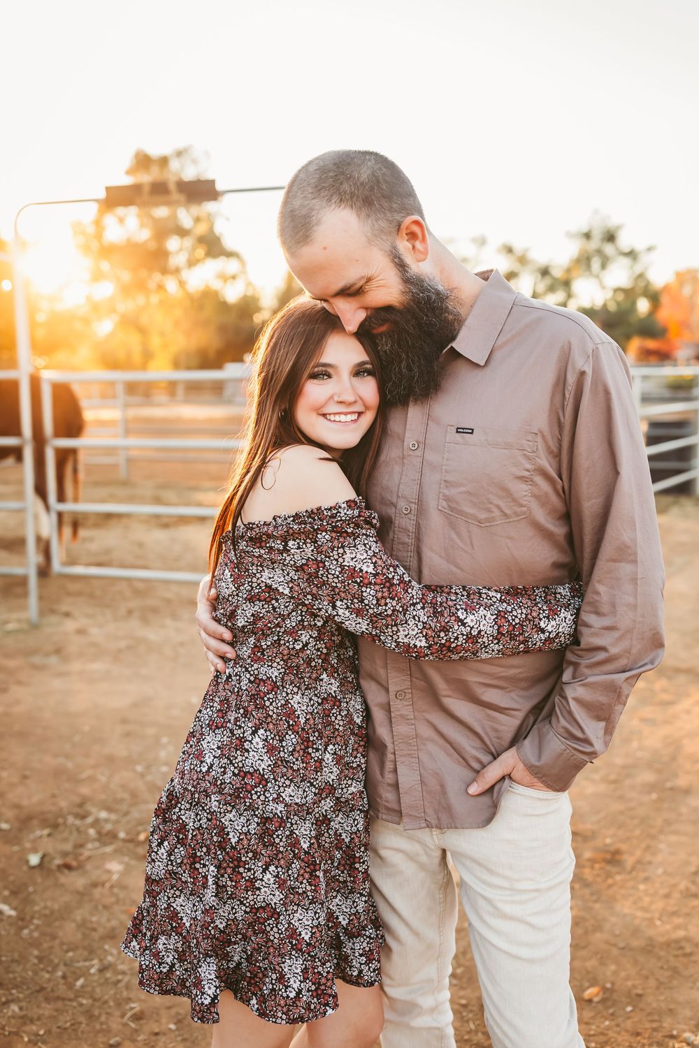 Couple embracing at a farm during golden hour, woman in floral dress smiling.