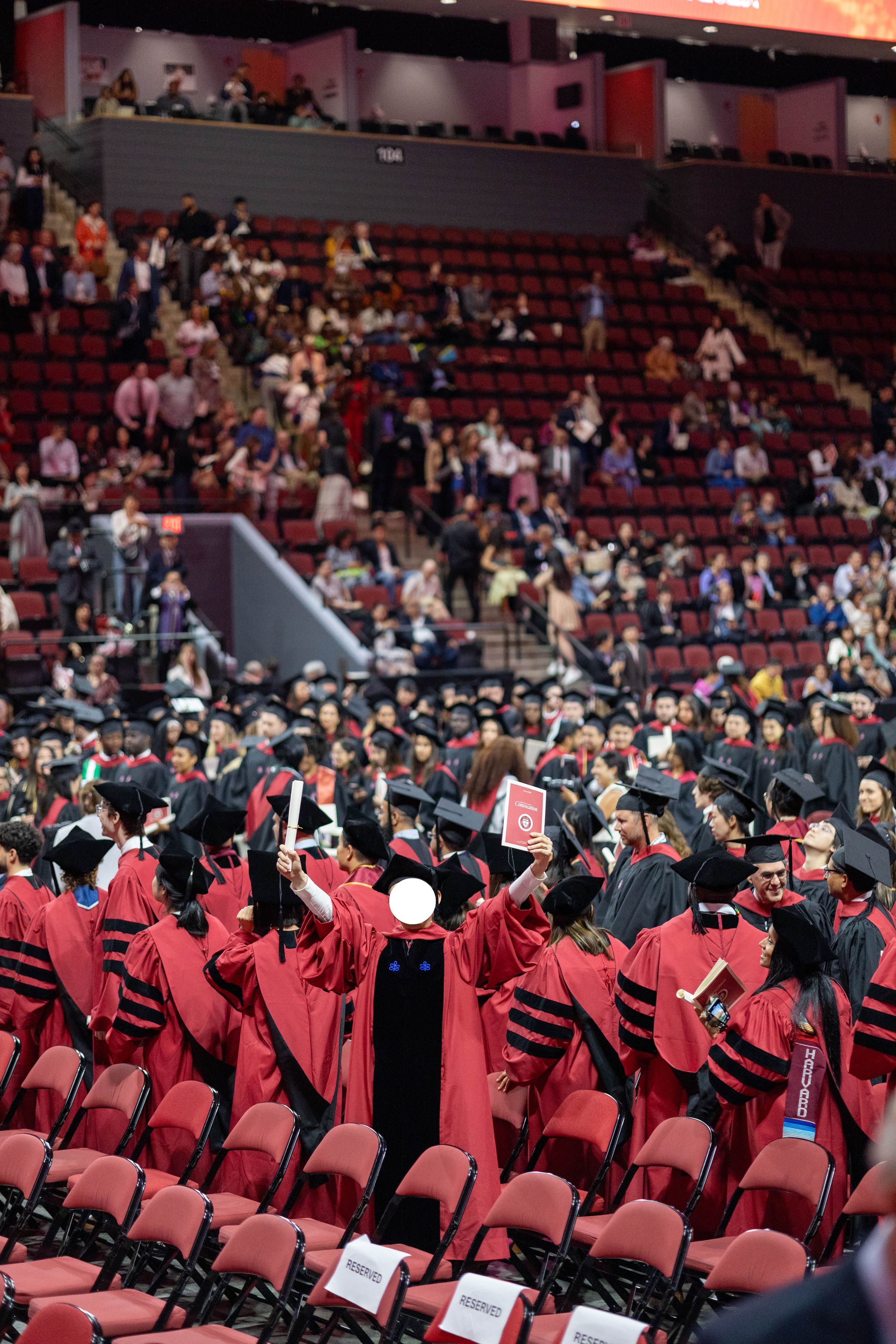 Graduates in red doctoral robes and black caps celebrate at a university commencement ceremony in a large arena.