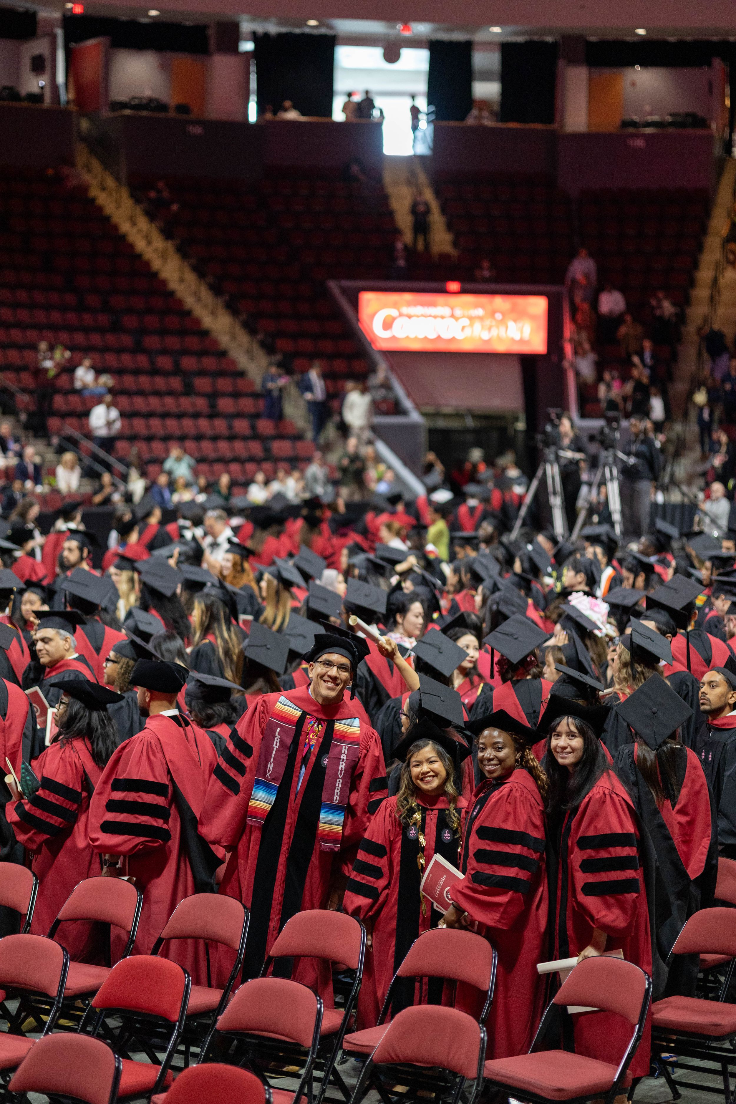 Graduates in red doctoral robes and black caps celebrate at a university commencement ceremony inside a large arena.