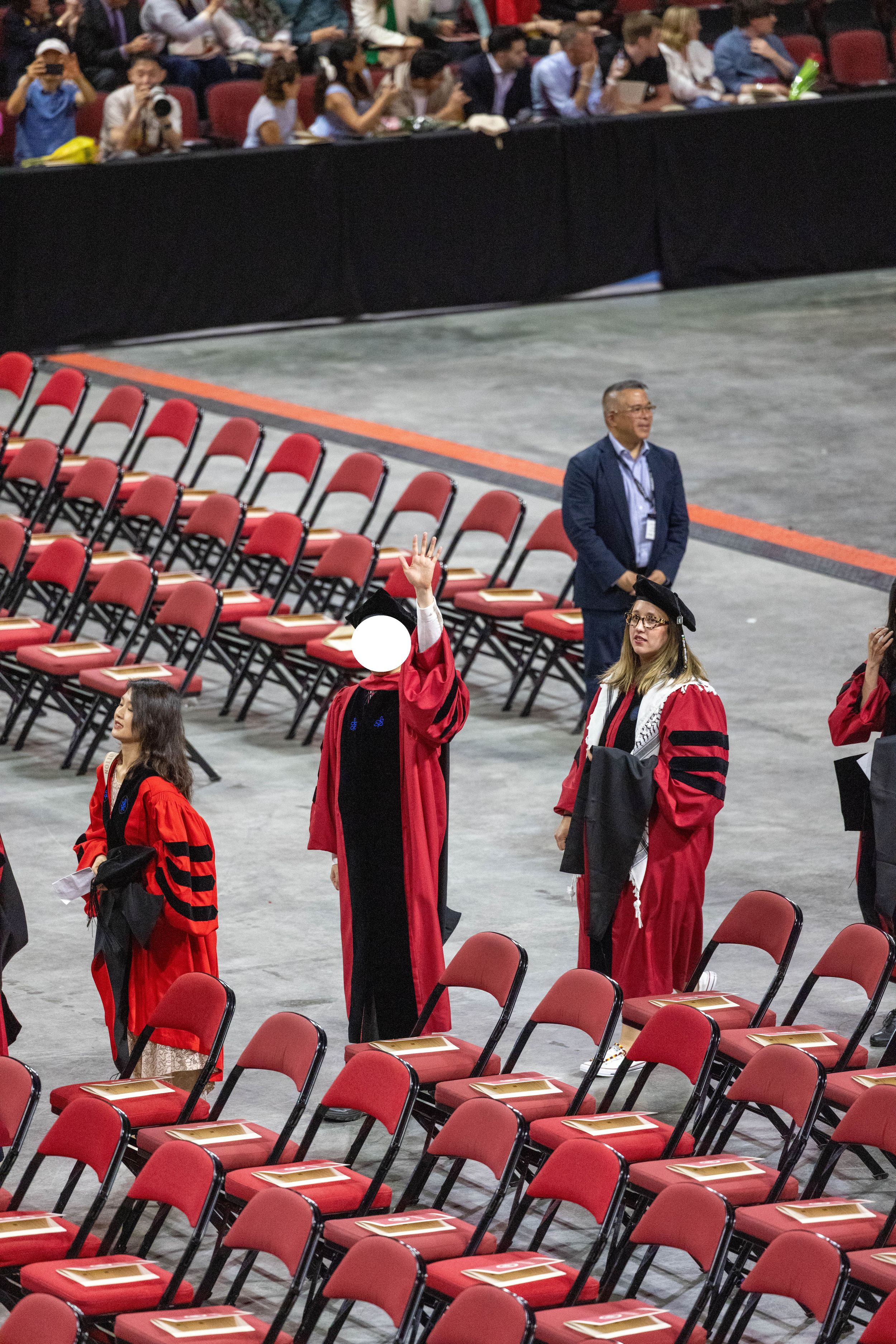 Graduates in red academic robes stand among rows of red chairs at a university commencement ceremony indoors.