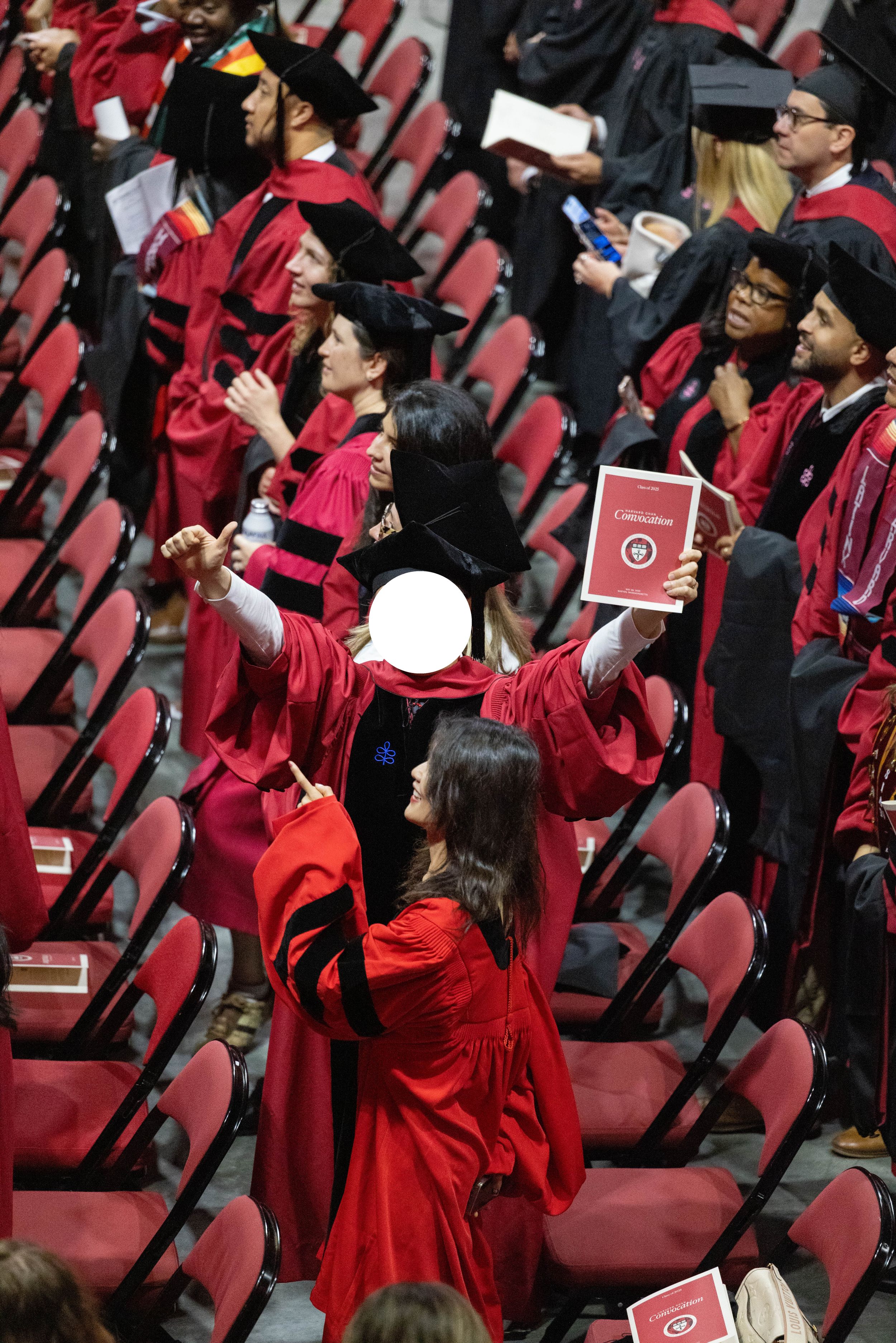 Graduates in red robes celebrate at a university commencement ceremony, seated in red chairs at an indoor arena.