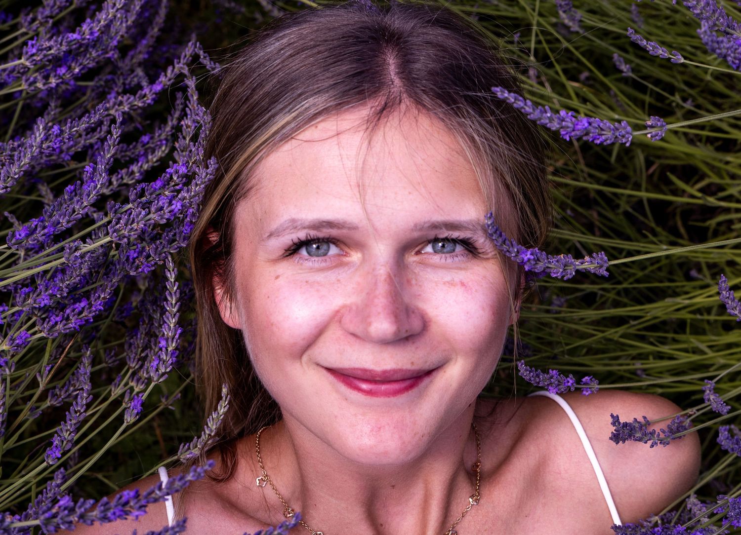 Young woman with freckles smiling among purple lavender flowers in a beautiful outdoor portrait.
