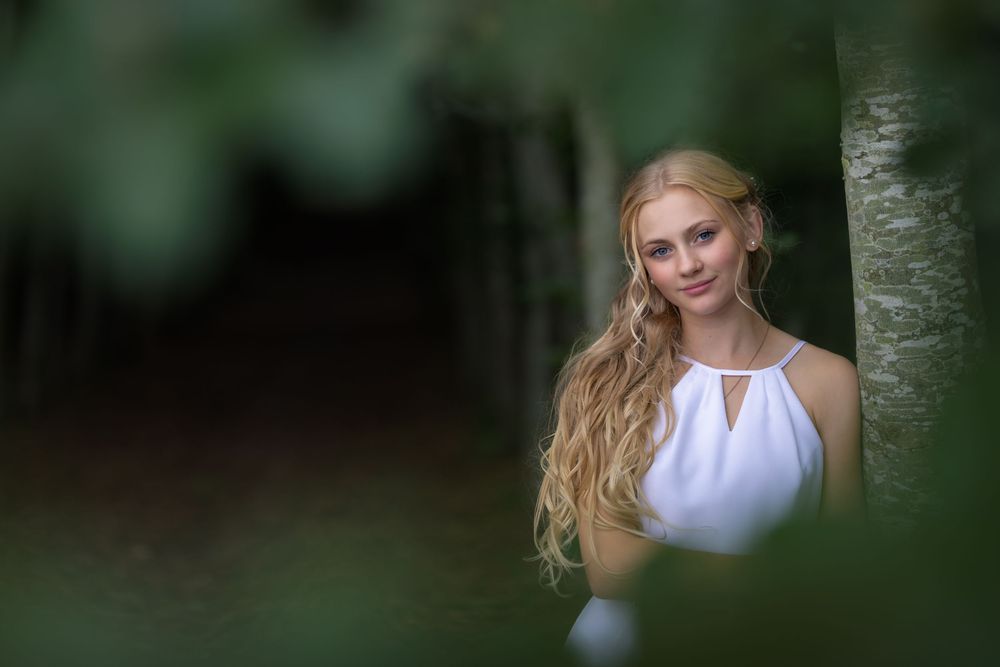 Young blonde woman in white dress posing gracefully among green trees in a serene forest setting.