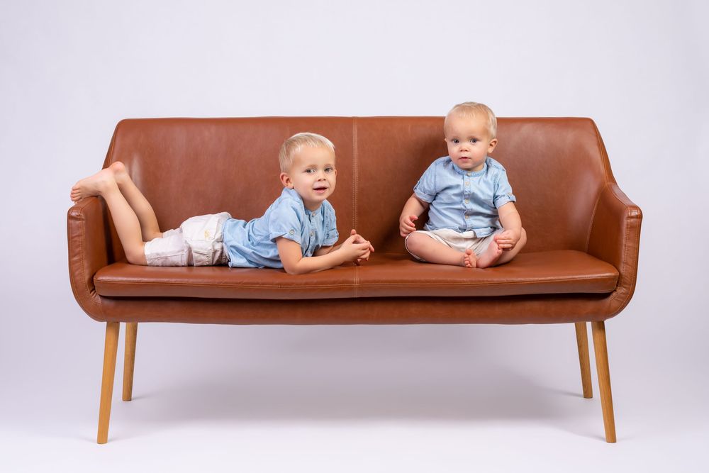 Two toddlers in matching blue shirts sitting on a brown leather couch against a white background.