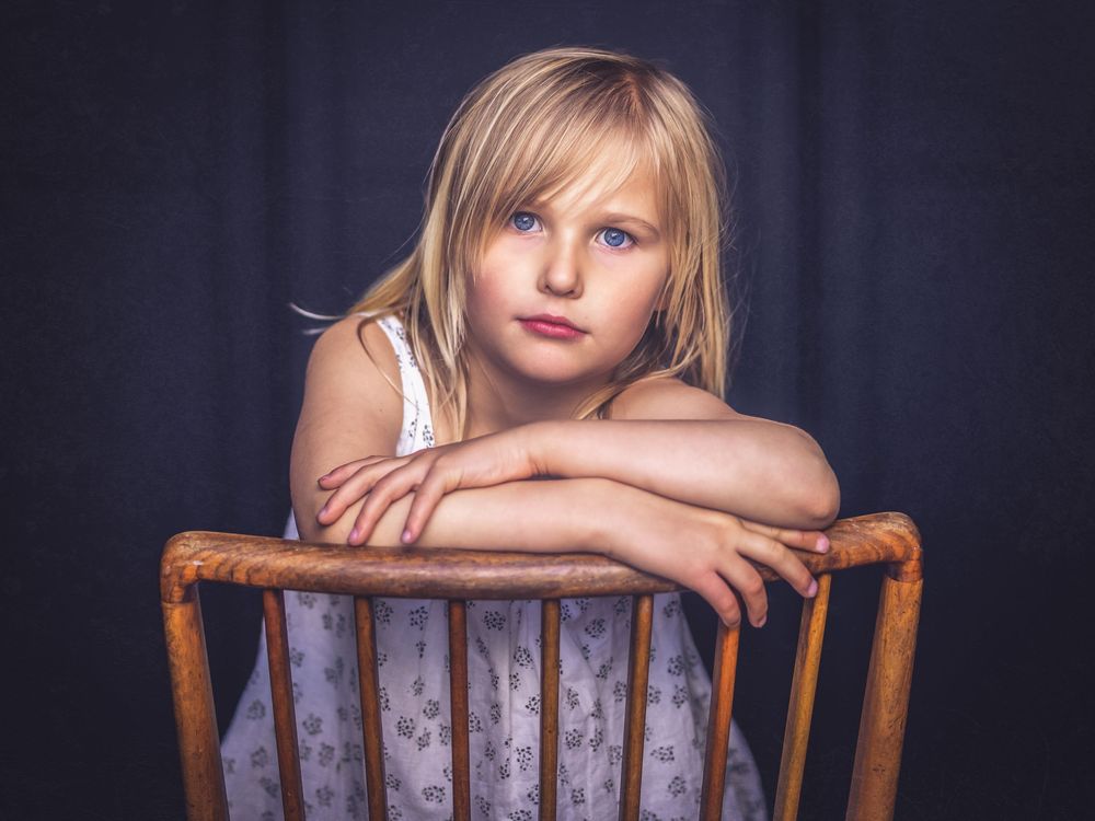 Young blonde girl with blue eyes resting arms on wooden chair back, posing for portrait against dark background.