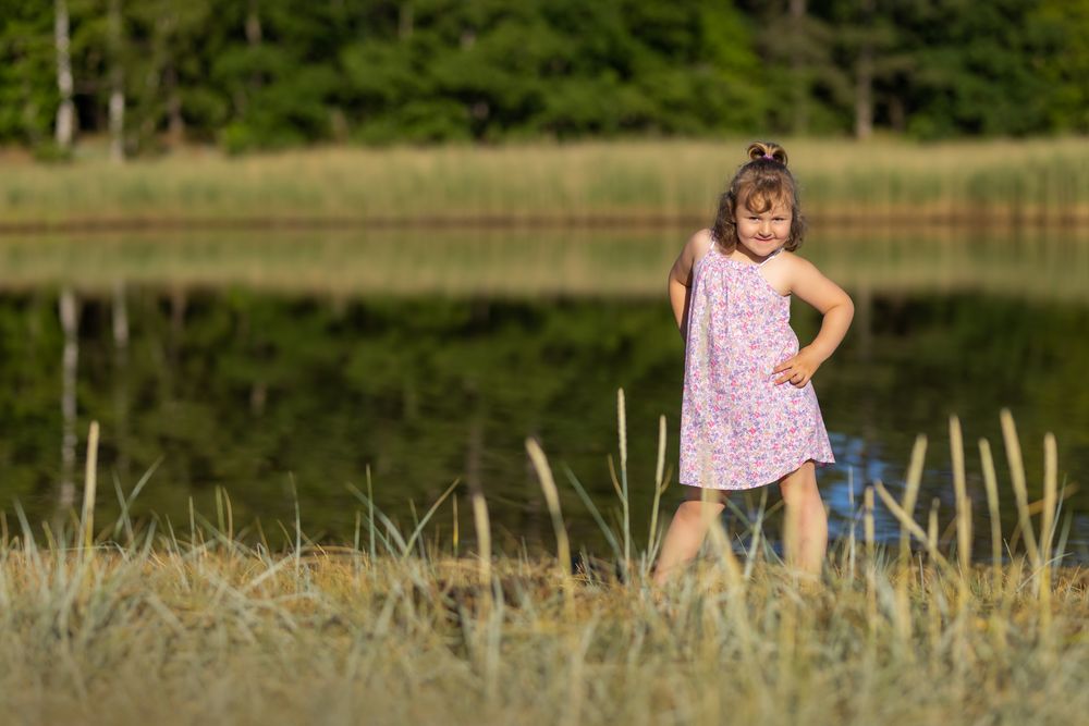 A young girl in a pink floral dress stands confidently by a lake in a grassy field.
