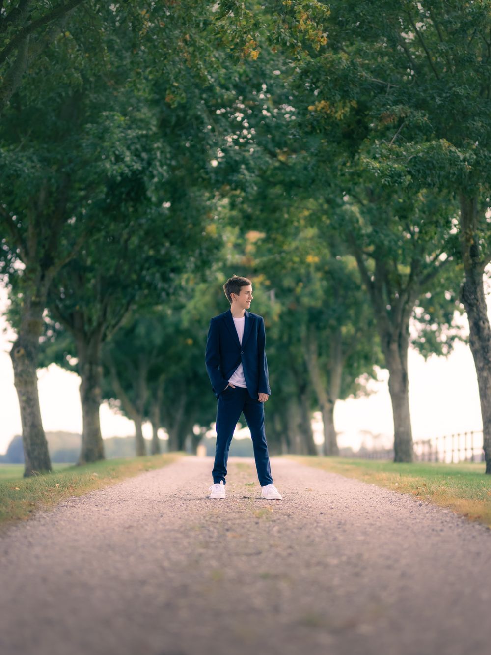 Young man in navy blazer and jeans standing on a tree-lined gravel path outdoors.
