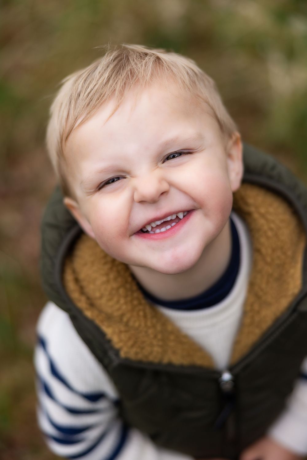 A cheerful toddler boy with blonde hair smiles broadly, wearing a striped shirt and green sherpa vest outdoors.