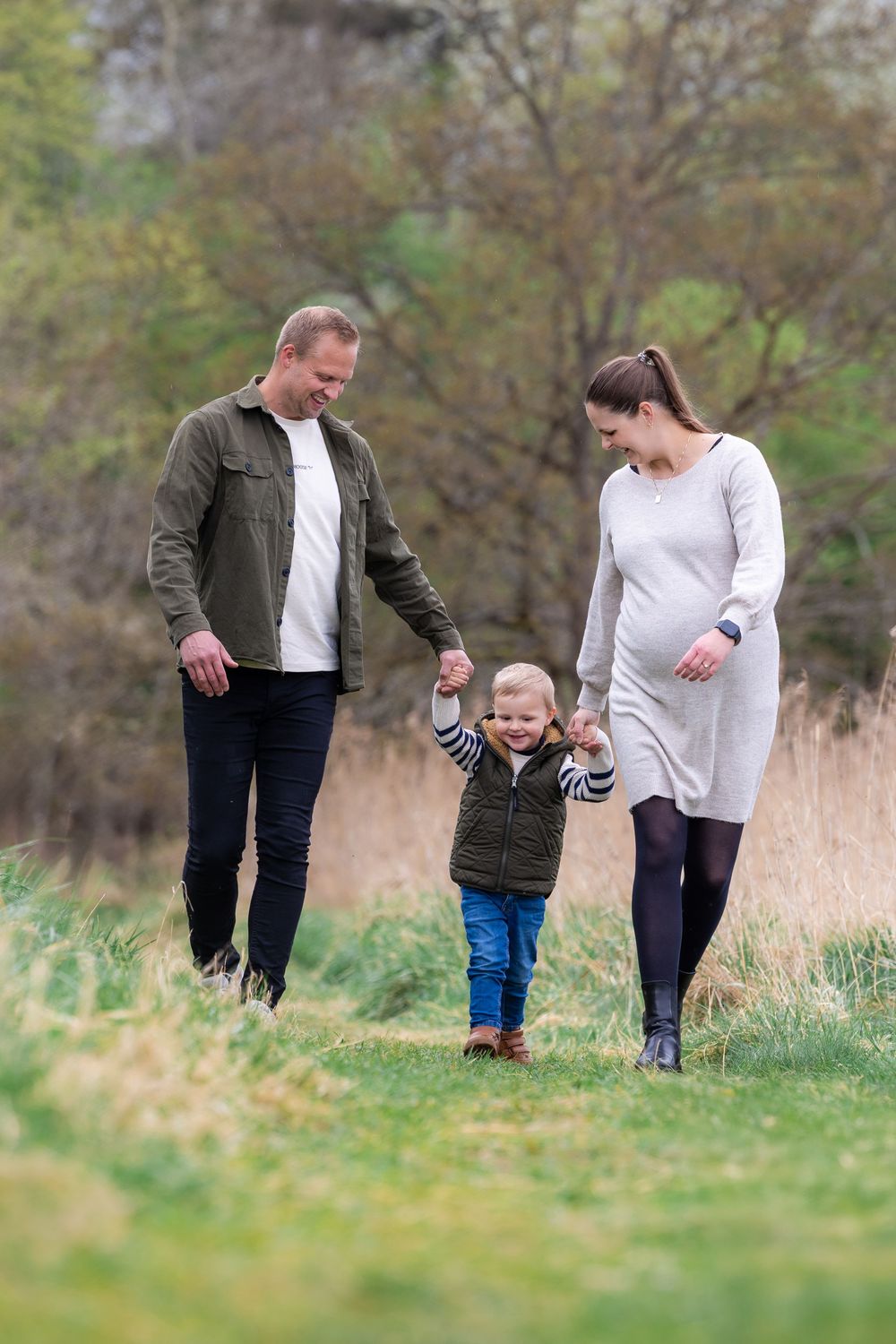 Pregnant woman and man walk hand-in-hand with toddler boy in green field surrounded by autumn trees.