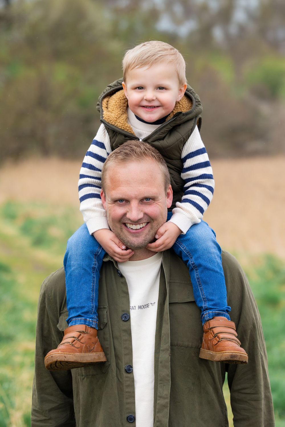 Smiling father carries happy toddler boy on his shoulders outdoors in a green field setting.
