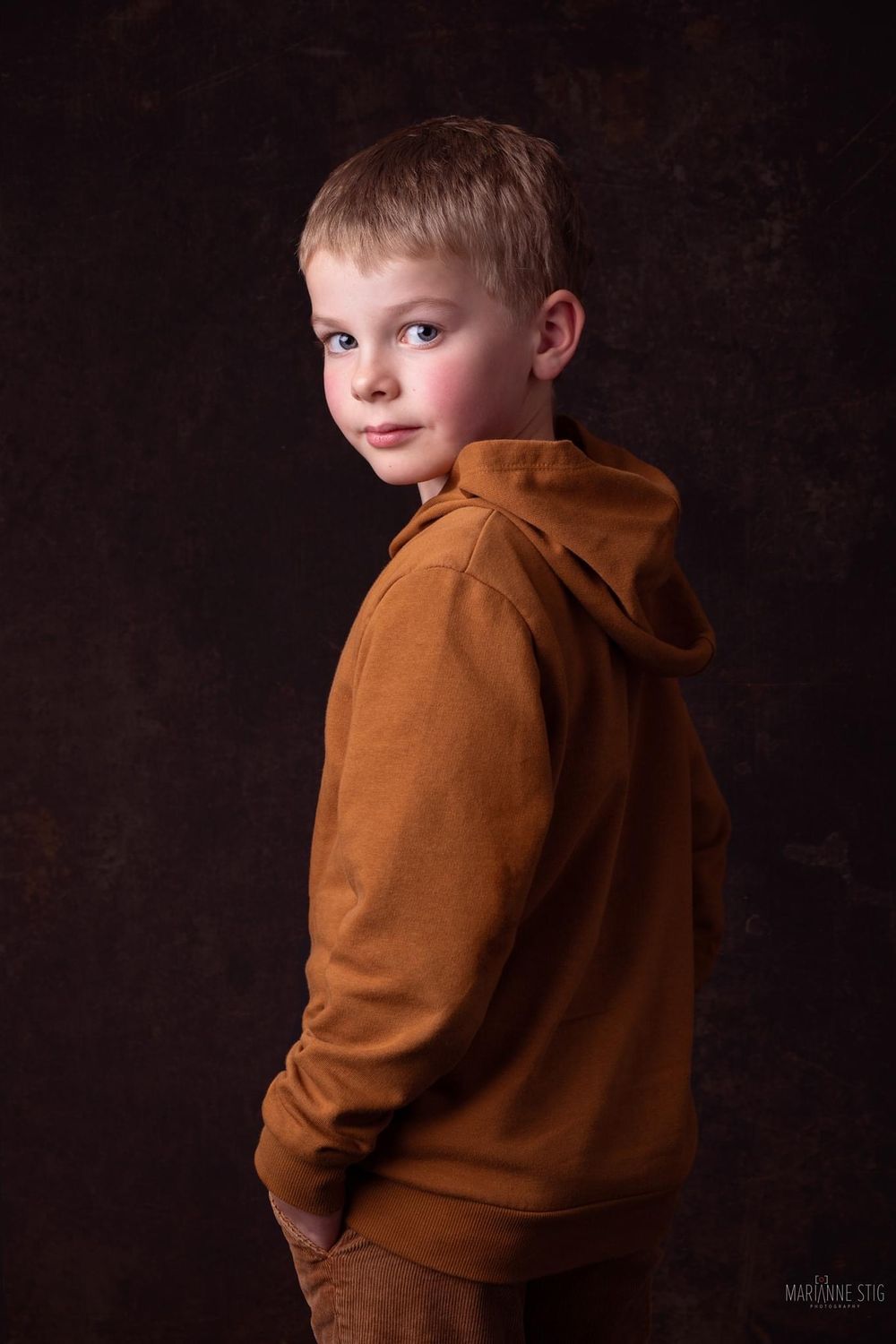 A young boy in a brown hoodie poses confidently against a dark studio background, glancing over his shoulder.