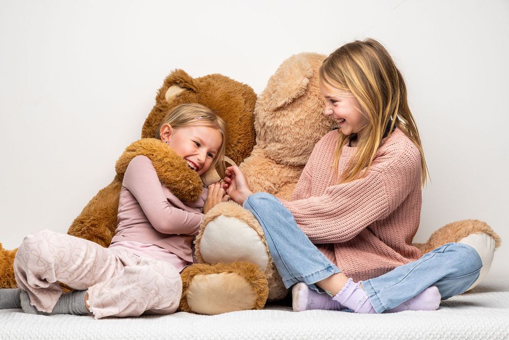 Two young girls sitting on floor laughing together with large brown teddy bears against white background.