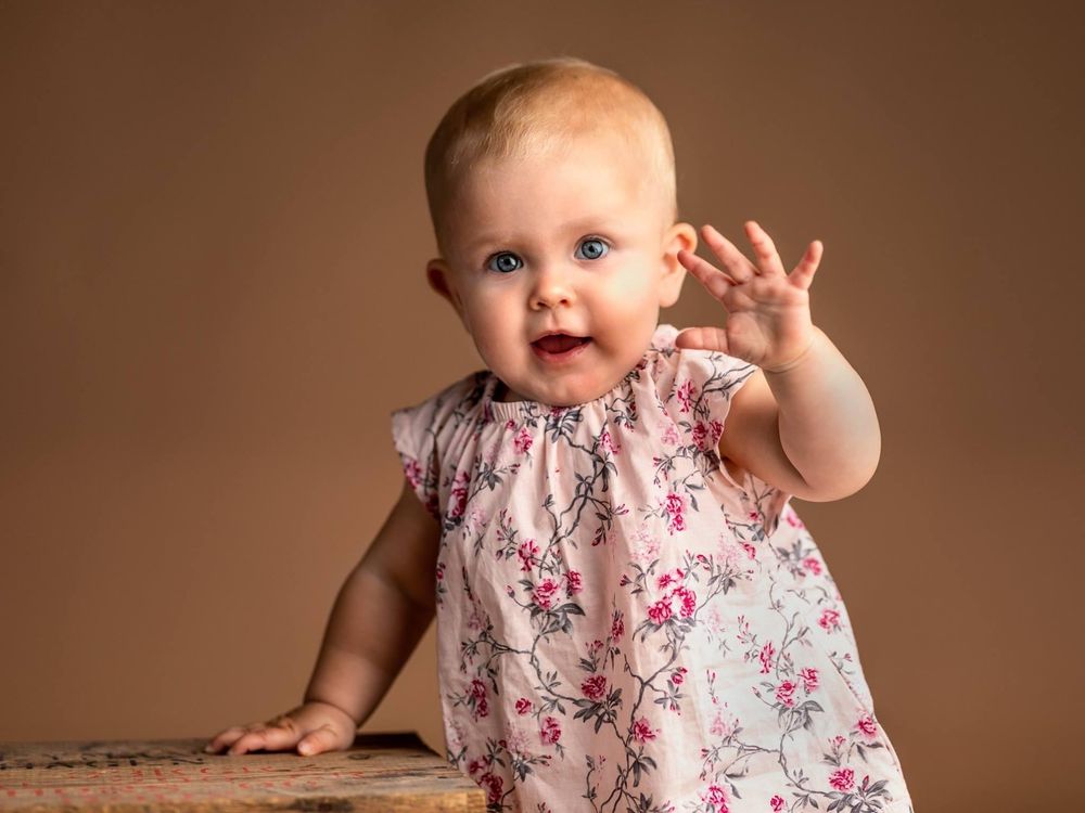 Adorable baby girl in floral pink dress with blue eyes waving at camera against brown studio background.