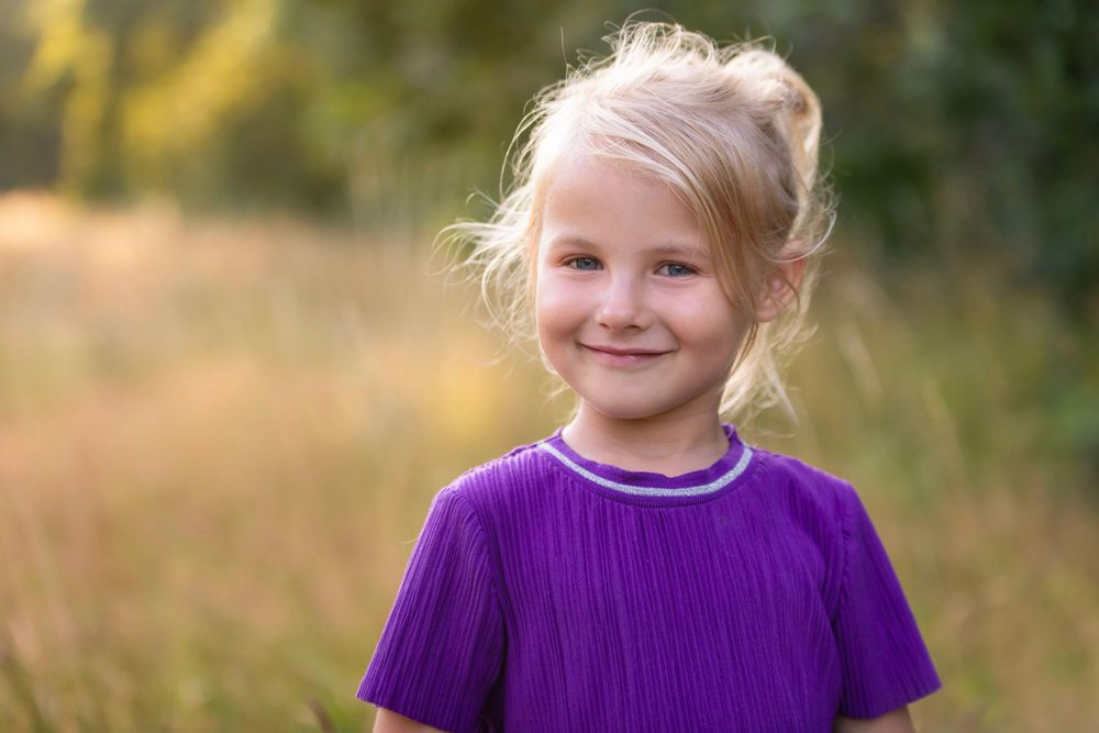 Smiling young blonde girl in a purple dress standing in a sunlit field with green trees in the background.