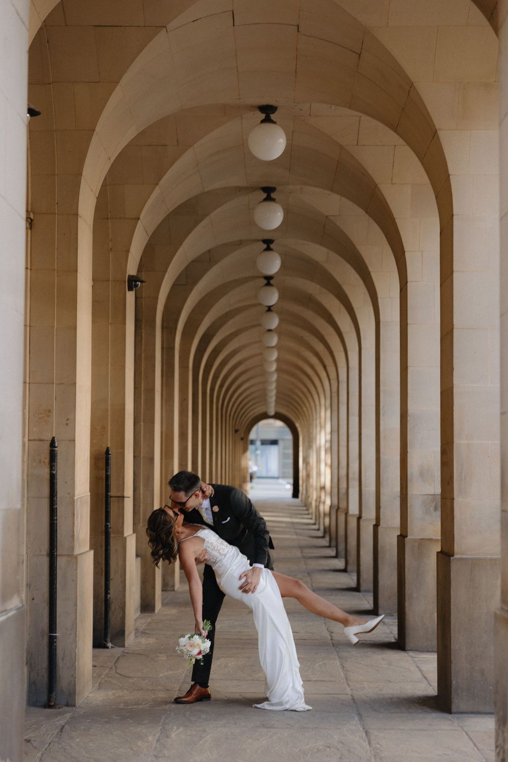 Bride and groom share a romantic dip kiss under elegant stone arches in a stunning wedding photo.