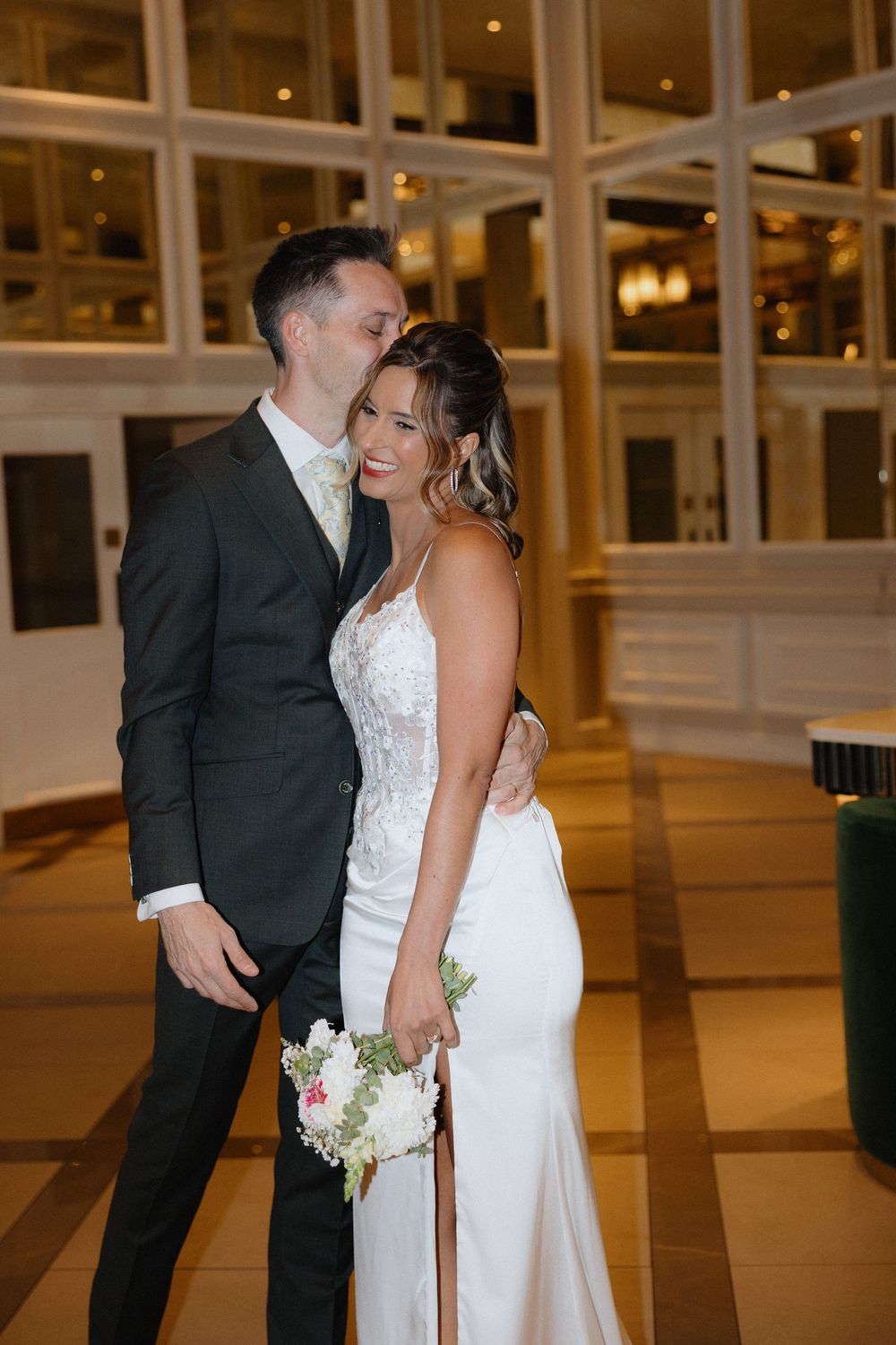Bride and groom share a romantic kiss pose in elegant wedding attire inside a beautifully lit venue.
