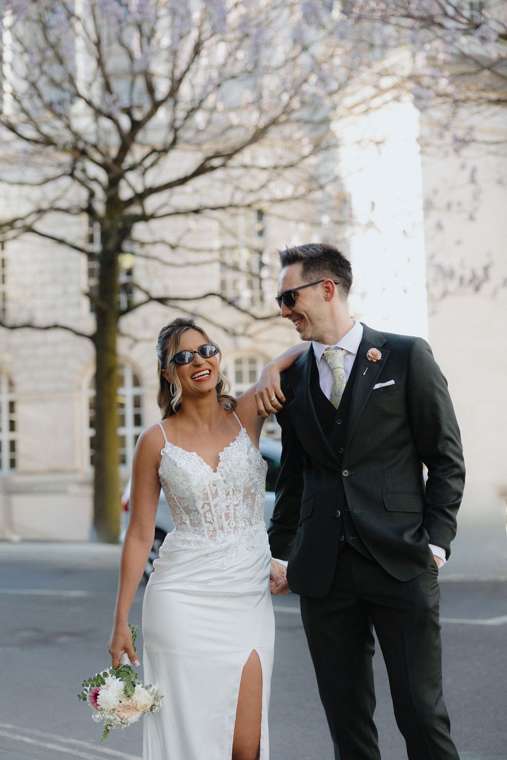 Smiling bride and groom in sunglasses walk hand in hand on a sunny city street, bride holding a bouquet.