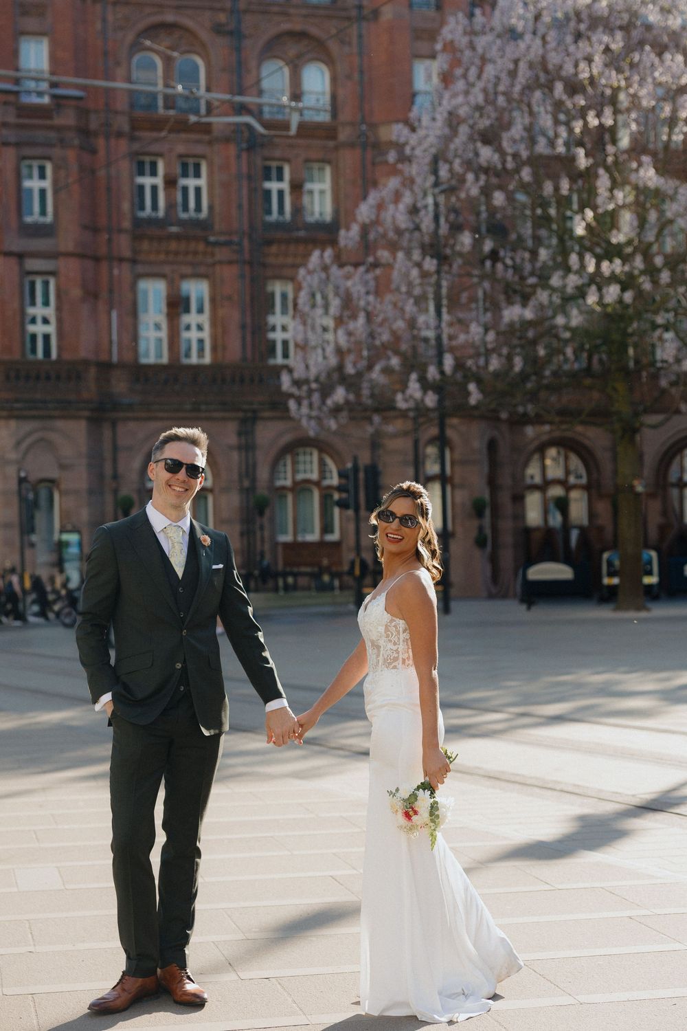 Bride and groom in sunglasses hold hands in a sunny city square with a blooming cherry tree behind them.