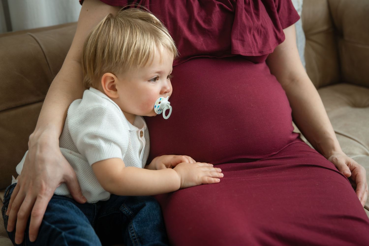 Toddler with pacifier touching pregnant mother's belly while sitting on a couch in a cozy home setting.