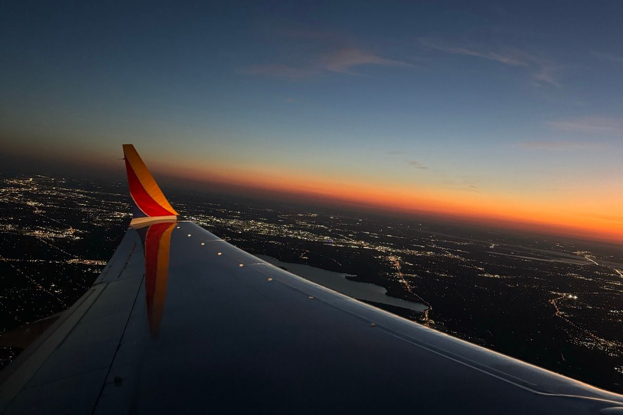 Southwest Airlines wing at sunset with city lights below and vibrant orange horizon during flight captured by The Mamones on travel.