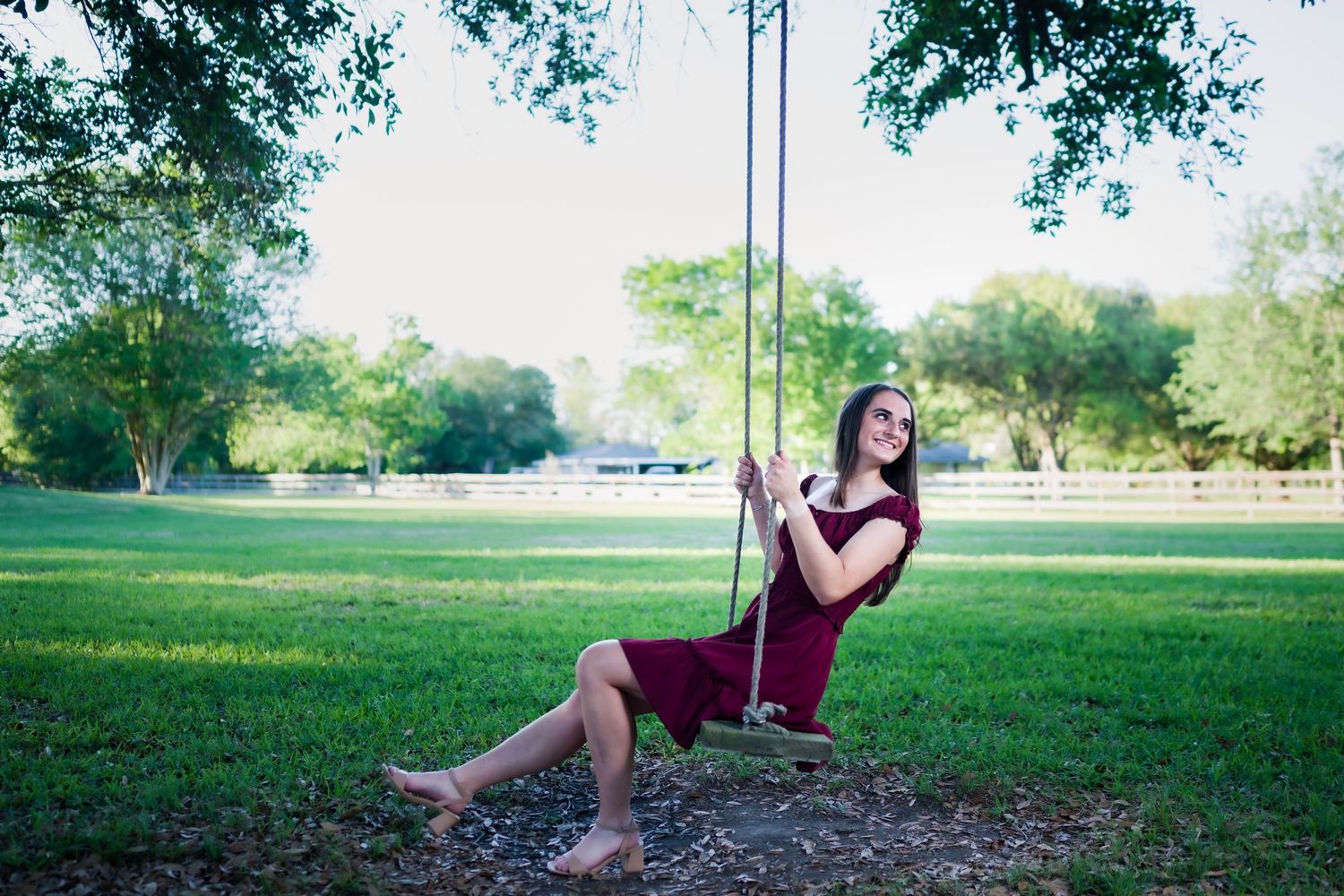 Young woman in burgundy dress sitting barefoot on a rope swing in a lush green park on a sunny day.