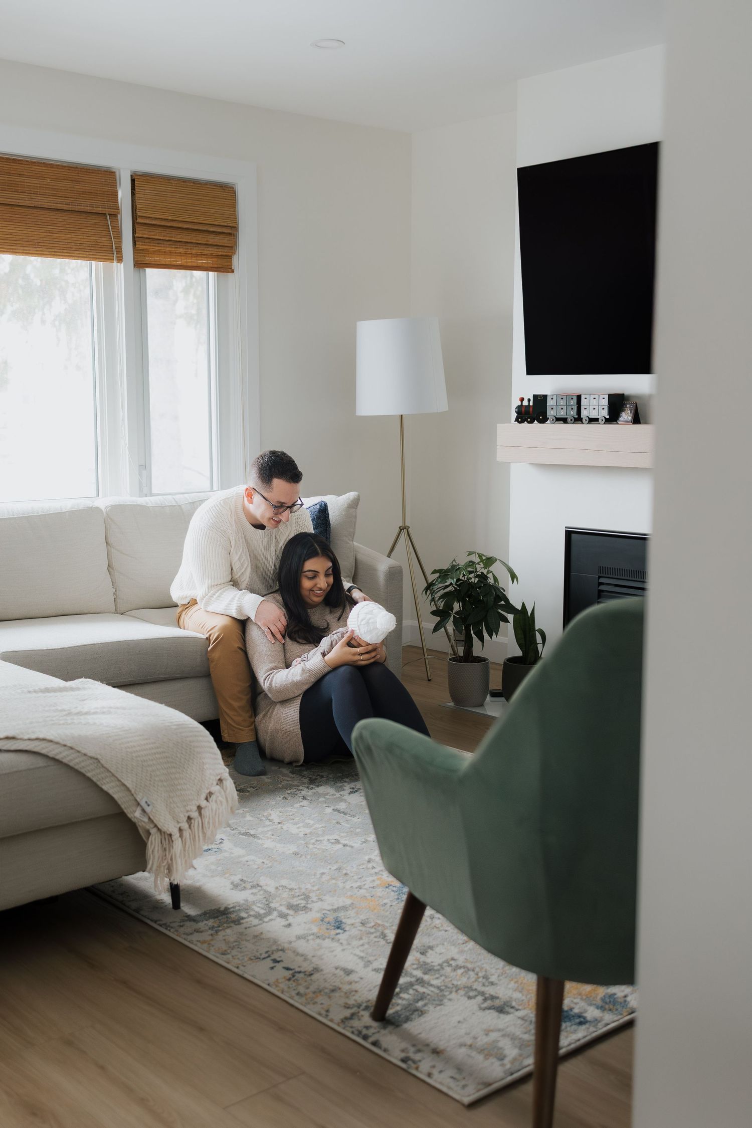 Parents in cream sweaters sitting on floor lovingly looking at newborn baby in white knit hat.